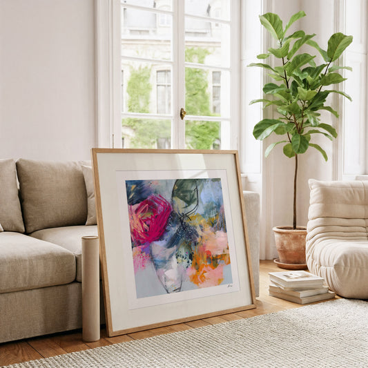 Framed abstract art piece on a wooden floor with a couch and plant in the background