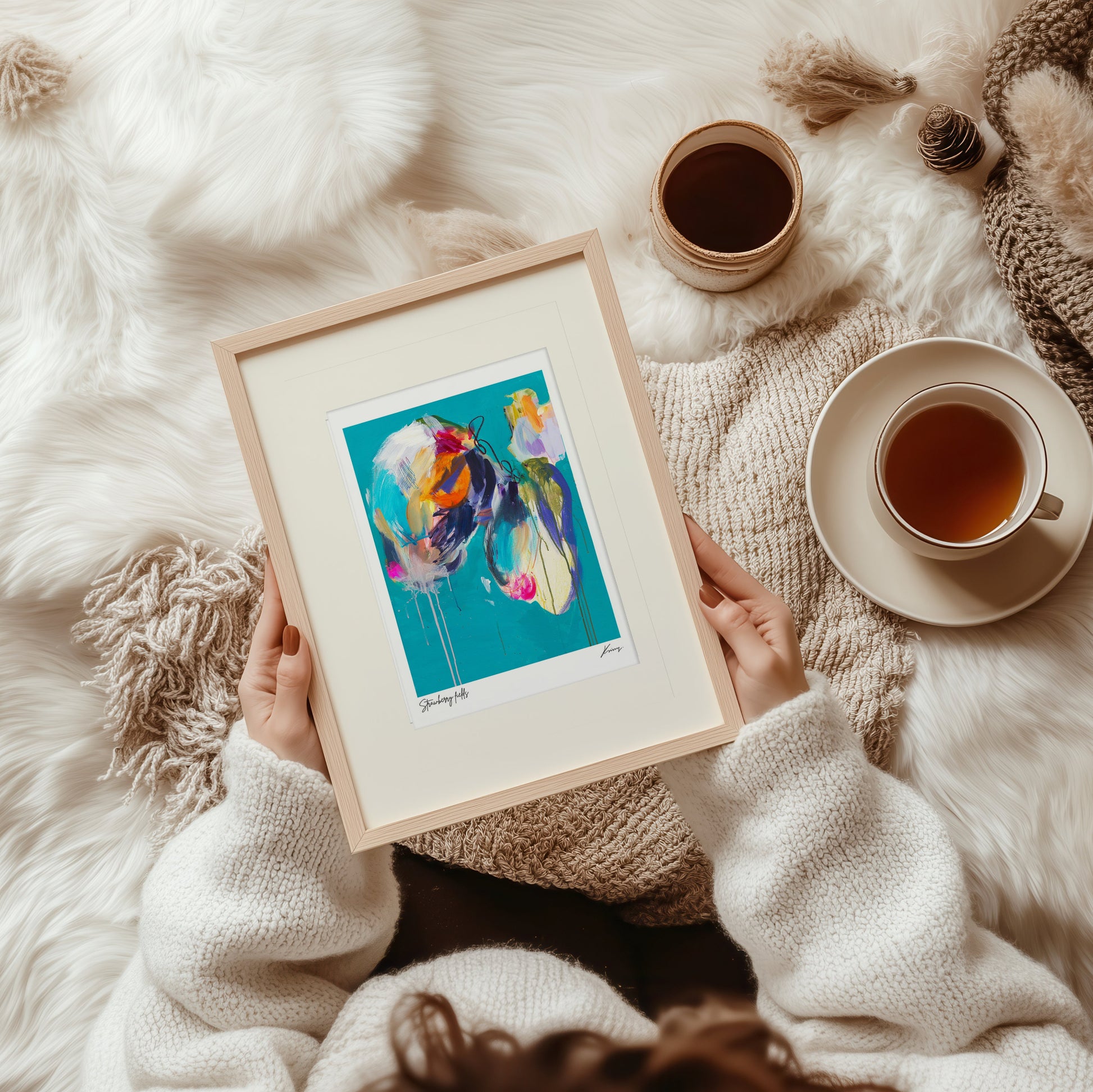 Person holding a framed colorful painting on a soft, white surface with a cup of tea and a knitted blanket.